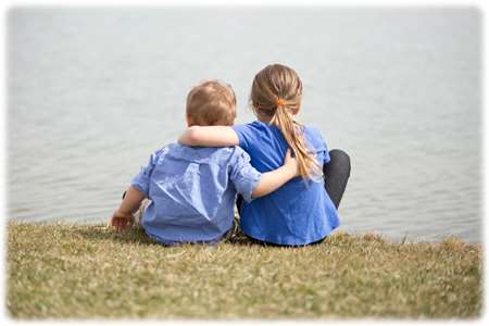 Image of a young brother and sister, from the back, sitting by a lake